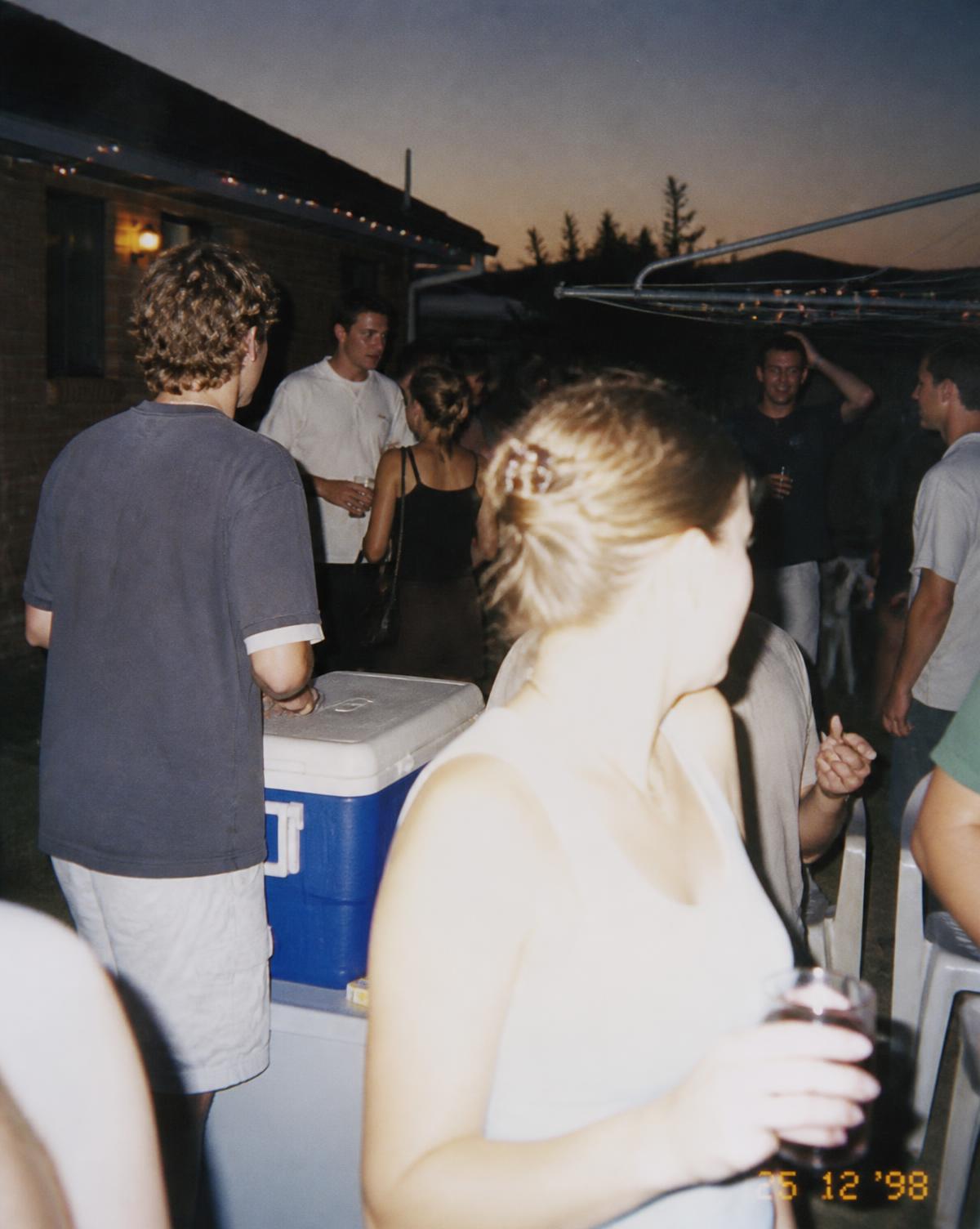 A late-1990s Australian backyard house party at summer dusk shot on a disposable camera with flash